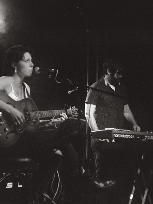 A black-and-white photo of two casually-dressed musicians: a young woman playing an acoustic guitar and singing into a mic, while a young man with a thick beard plays a synthesizer a few steps away.