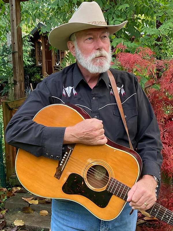 A ruddy-faced man with a full, neatly trimmed beard stands in front of a forested backdrop, staring determinedly out of frame. He wears a neat white cowboy hat and black button-up shirt with white trim and cow skull accents, and holds an acoustic guitar closely with both hands.