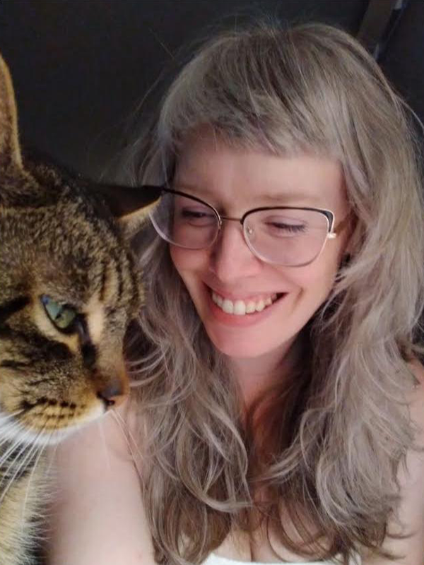 A young woman with horn-rimmed glasses and dark blonde hair smiles sweetly at her cat in the foreground.