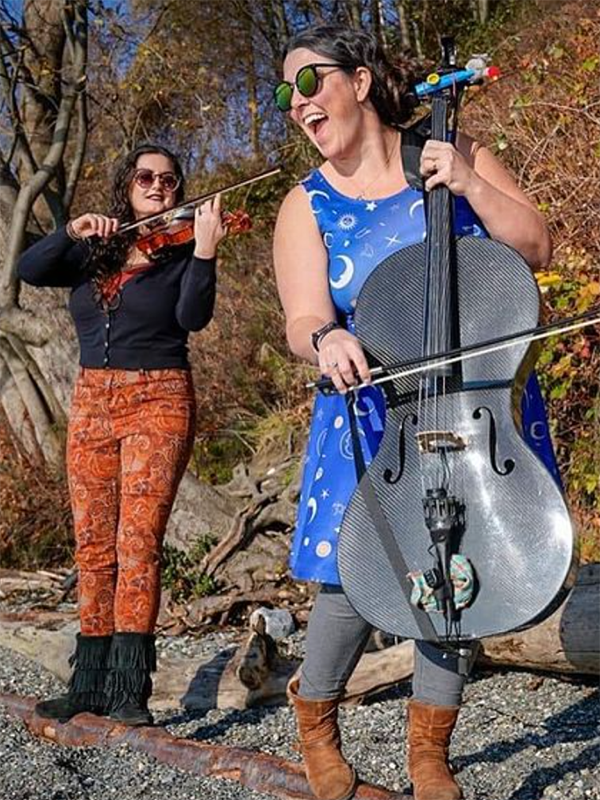 A colorful photo of two brunette women in sunglasses on a rocky Northwestern beach. One wears a blue dress with stars and moons and sings boisterously while playing a stand-up electric cello. The other, a few steps behind, wears funky orange pants and a black sweater, playing a fiddle.