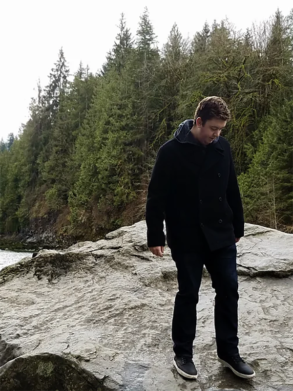 A young man pensively stands on a large rock span, staring downward at the stone. Behind him, a coniferous forest thickly borders a rushing river.