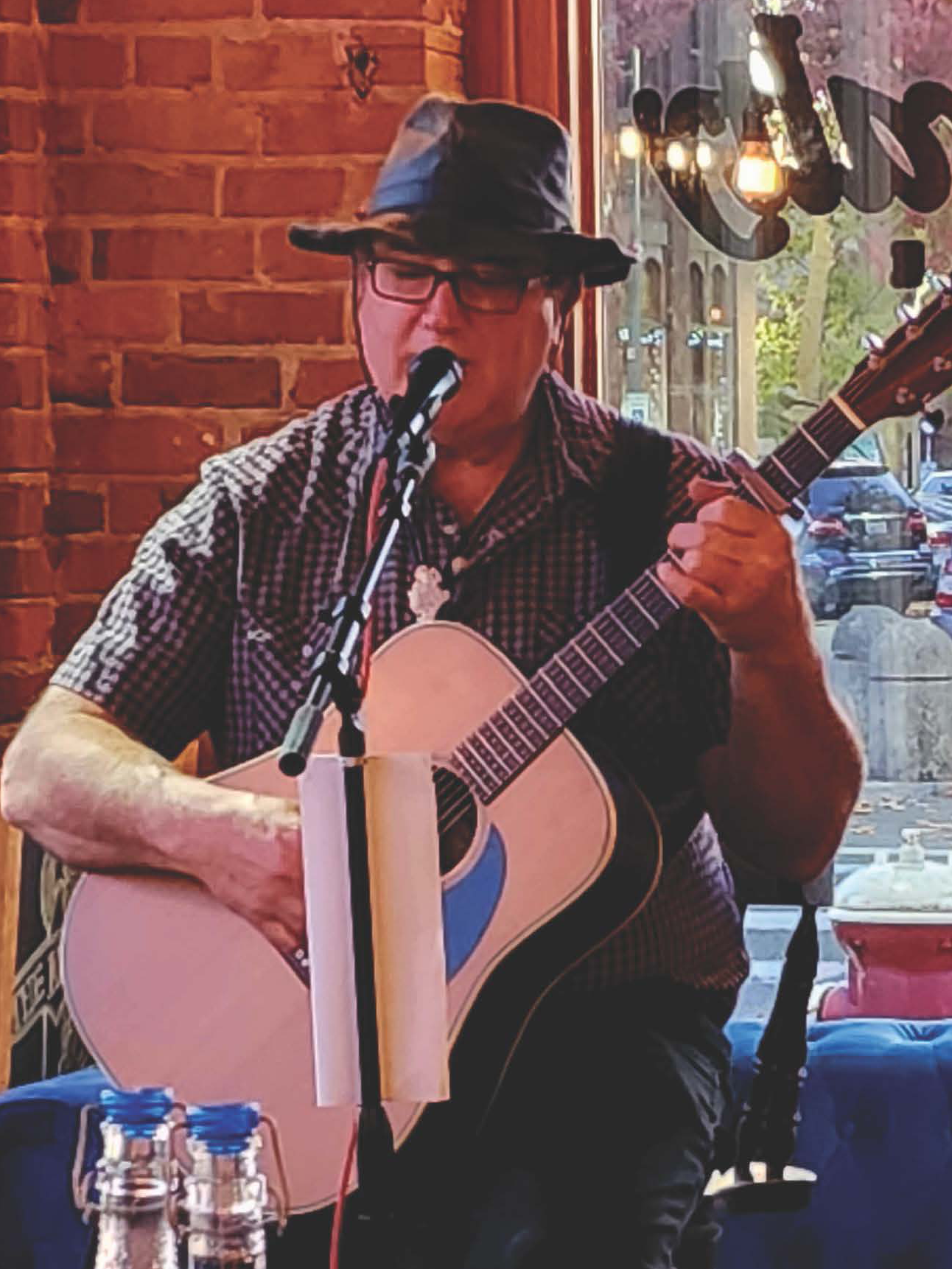 A 50-ish white man in spectacles, a plaid shirt, and a wide-brimmed hat sings into a microphone while playing an acoustic guitar.