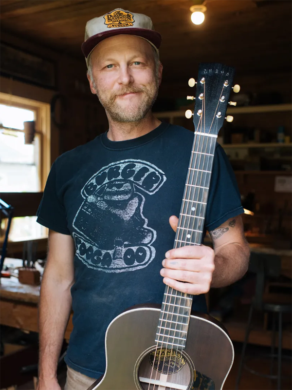 A 40ish white man with short-trimmed facial hair gives a thoughtful, friendly smile to the camera. He wears a trucker cap and a faded t-shirt and holds an acoustic guitar in one hand.
