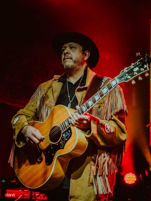 A 50-ish Native man with thick stubble and a small mustache plays an ornate acoustic guitar while staring intently into the distance. Stage lights shine behind him, suggesting that he is on a stage. He wears a wide-brimmed black hat and a brown leather jacket with plenty of fringe.