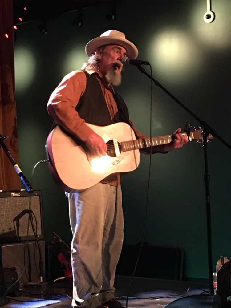 A mature-looking older man with a long, white beard and folksy garb plays an acoustic guitar and sings into a microphone on stage.