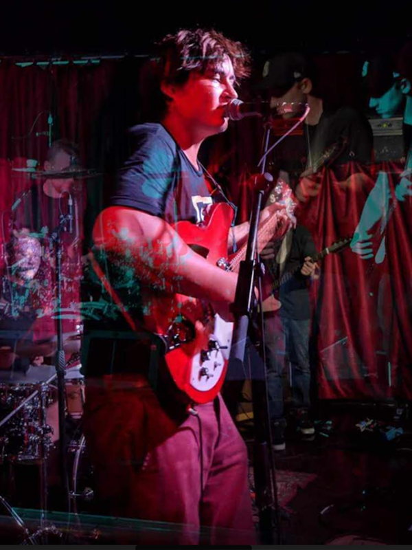 A young man plays an electric guitar while singing intently into a microphone. A drummer and a guitarist play behind him in a psychedelically shifting and colorful background.