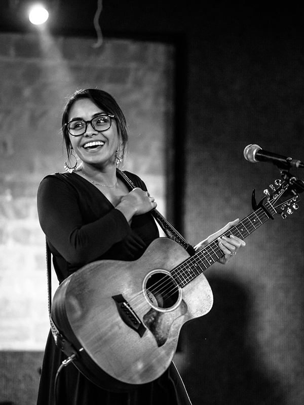 A Mexican-American woman with straight hair and glasses flashes a brilliant smile during a concert. She holds a guitar and stands between a stage light and a microphone.
