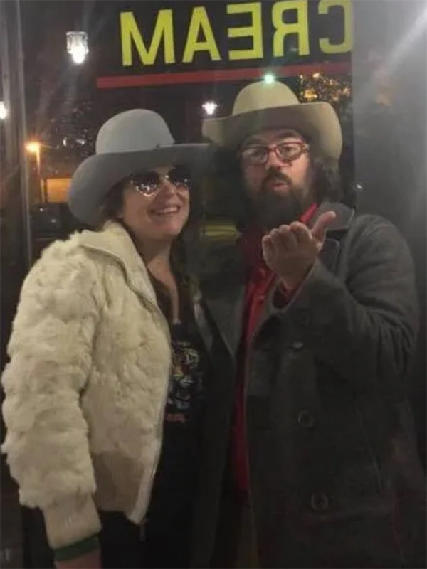 Two young white folks in beige cowboy hats and big jackets smile and blow a kiss toward the camera.